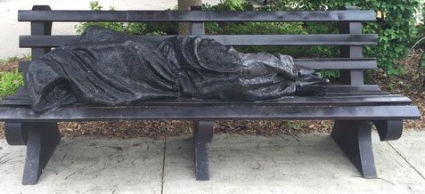A statue of a person sleeping on a bench outside.